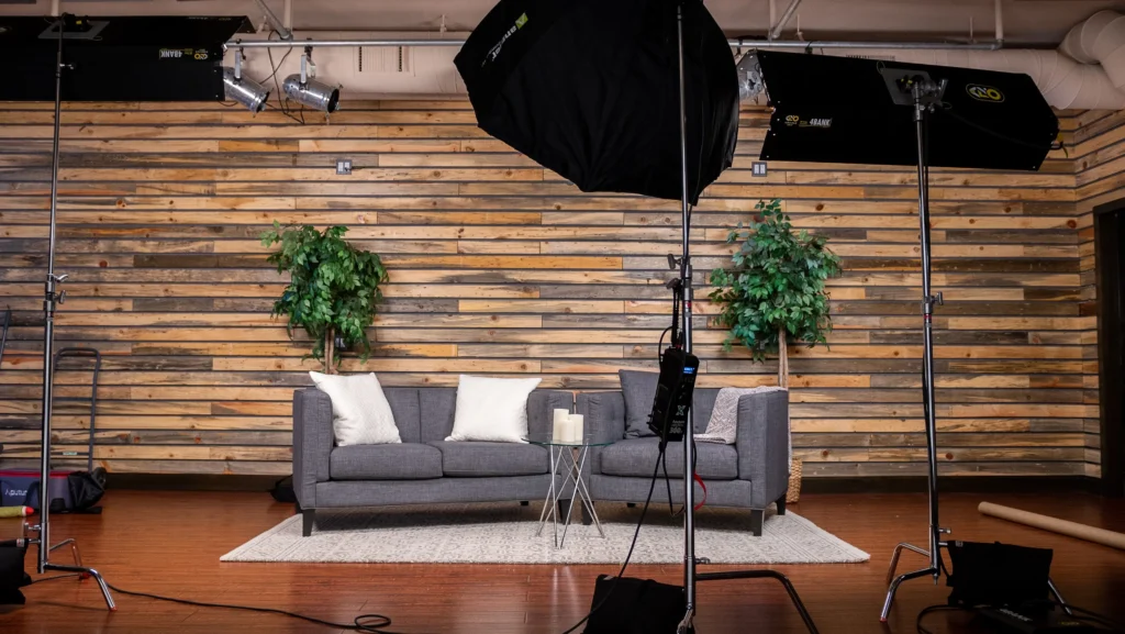 Sound-Stage-Gallery-16 - Hermosa Sound Lab A professional video or photo studio setup with a gray couch, two green plants, lighting equipment, and a camera on a tripod.